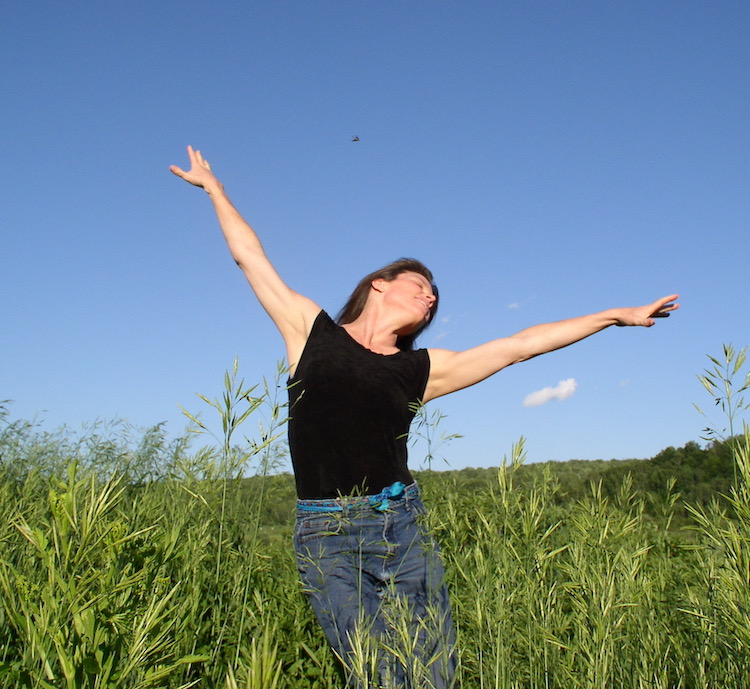 a woman in jeans and a tight black top dances against a clear blue sky in a green field, she has fair skin and dark hair and her arms are outstreched ecstatically